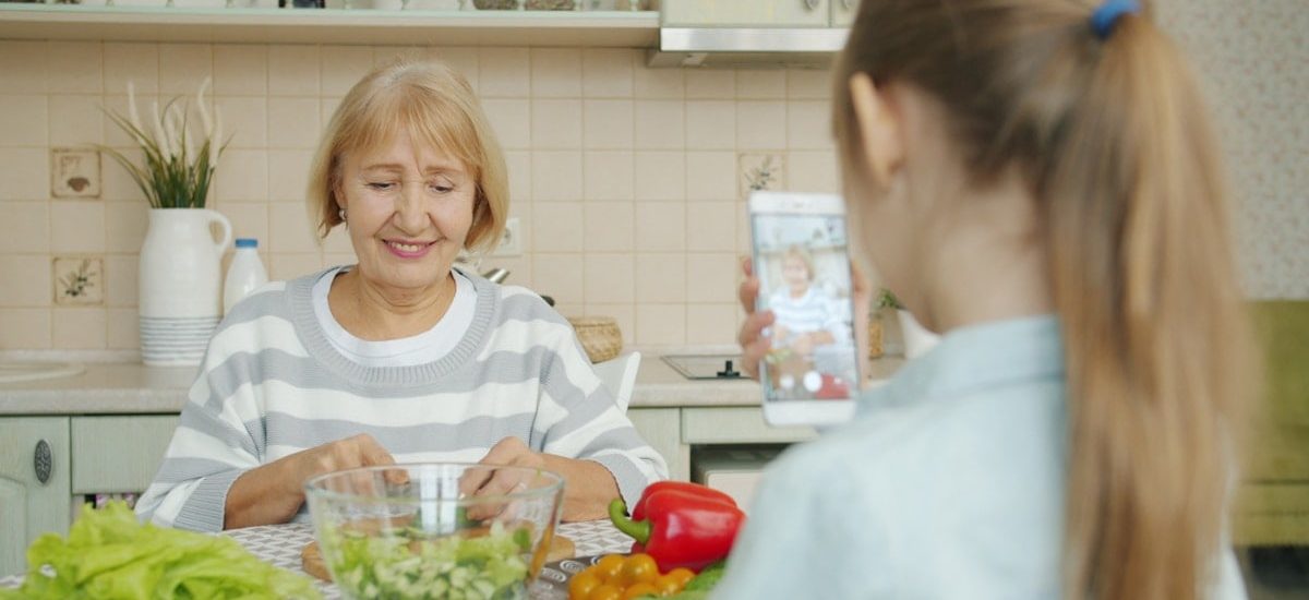 Cheerful elderly woman is posing for smartphone camera and cooking salad while child is taking picture with modern device. Family, photograph and people concept.