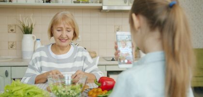 Cheerful elderly woman is posing for smartphone camera and cooking salad while child is taking picture with modern device. Family, photograph and people concept.