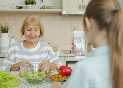 Cheerful elderly woman is posing for smartphone camera and cooking salad while child is taking picture with modern device. Family, photograph and people concept.